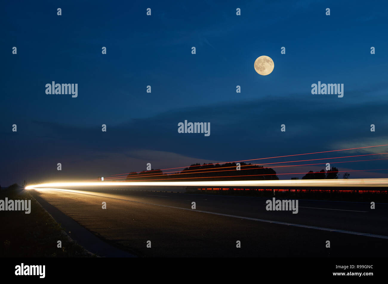 Luna piena sopra l'autostrada, fari di luce splendente linee in movimento le vetture nel paesaggio al crepuscolo. Blu scuro cielo estivo con le nuvole. Foto Stock