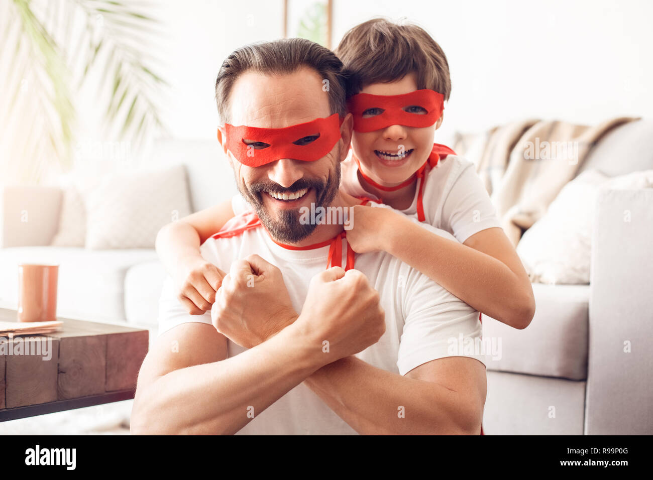 Padre e figlio indossando costumi superheroe insieme a casa abbracciando guardando la telecamera sorridendo felice team close-up Foto Stock