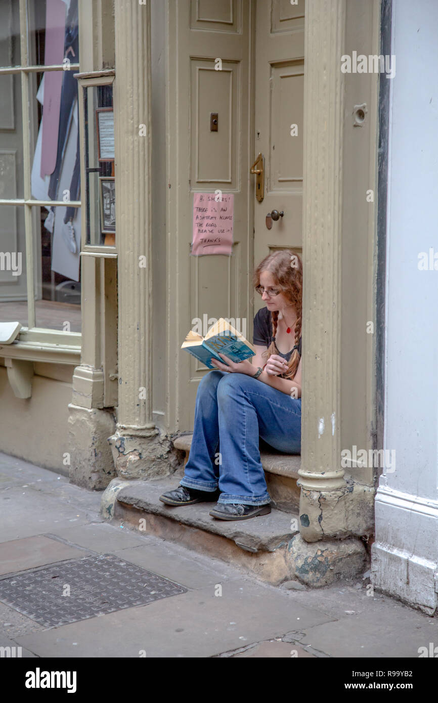 Una giovane donna che legge un libro in una porta a. un negozio mentre fuma durante la sua pausa di lavoro accanto a. avviso per dissuadere i fumatori Foto Stock