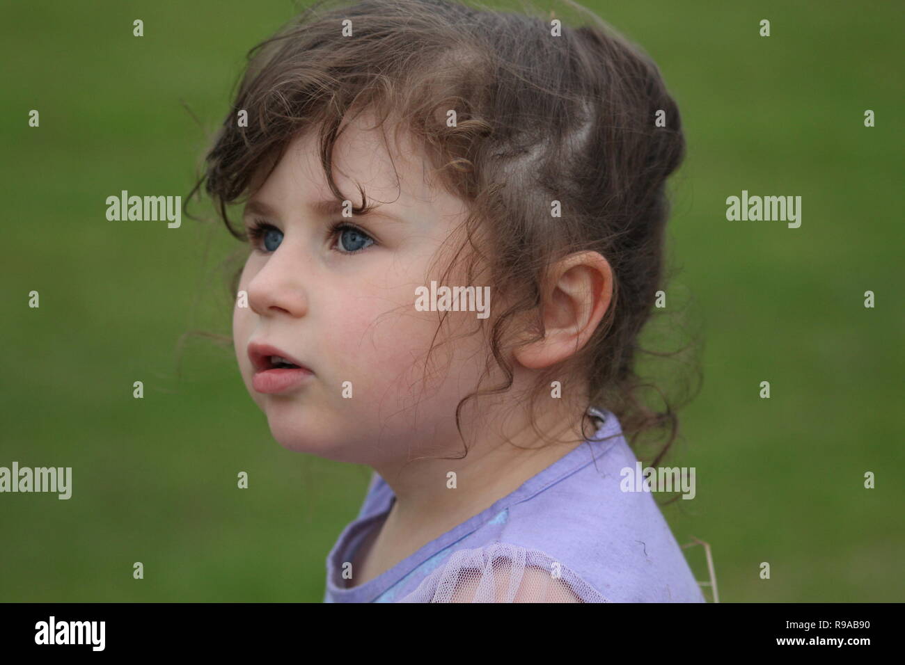 Giovane bruna bambino con grandi occhi blu che guardano lontano dalla macchina fotografica su uno sfondo di erba Foto Stock