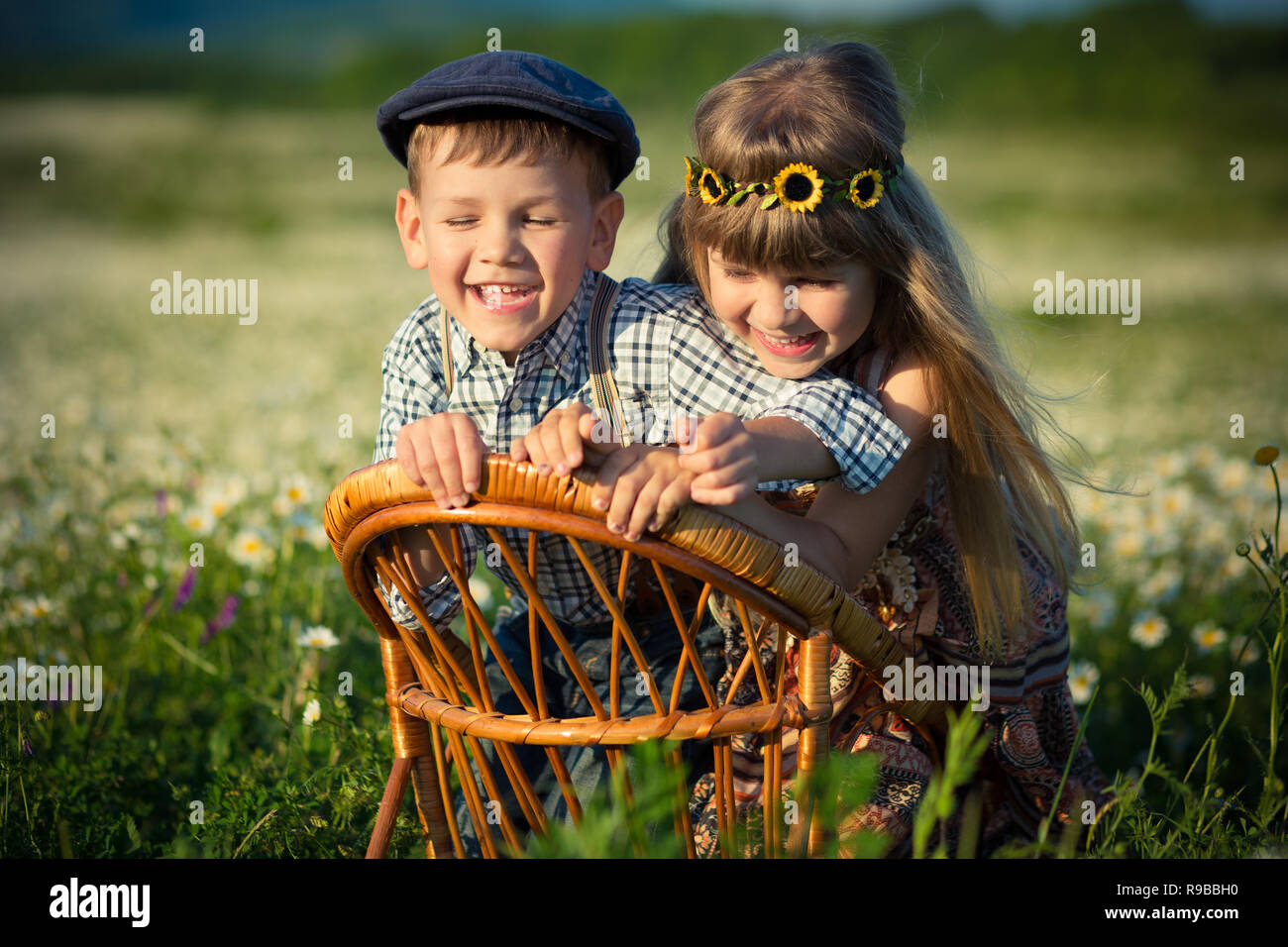 Carino bello ragazzo che indossa jeans e maglietta sittingand giocando sulla sedia di legno insieme con una bellissima ragazza sul prato di chamomiles daisy e sorridente e Foto Stock