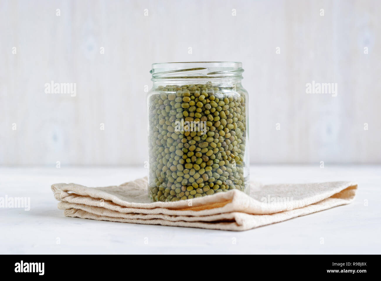 Memorizzazione vetro vaso pieno di verde secchi Mung Bean su un panno da cucina su uno sfondo bianco visto dal lato Foto Stock