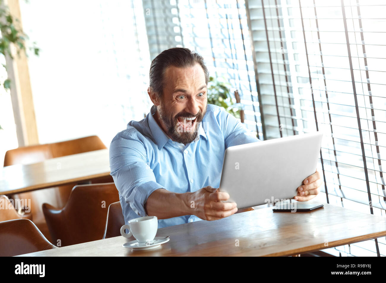 Uomo maturo trader seduta a tavola al cafe di giorno con tazza di caffè azienda laptop che stanno guardando sullo schermo grida eccitato Foto Stock