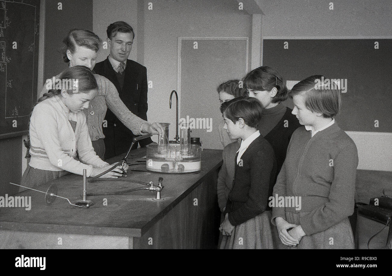 1955, una scolaretta facendo un esperimento su un banco da lavoro in una classe di scienze, aiutato da un insegnante e guardato dai suoi compagni di scuola, England, Regno Unito Foto Stock
