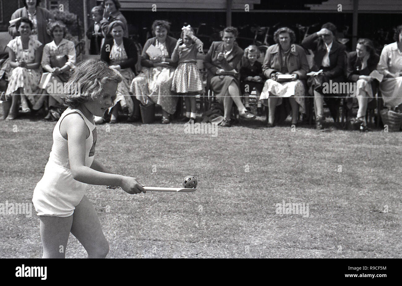 Degli anni Cinquanta, storico, scuola di sport, una giovane ragazza di indossare abbigliamento da palestra - shorts e un giubbotto - tenendo la palla con un cucchiaio di legno in un uovo e cucchiaio gara, guardato dagli alunni ai genitori, Inghilterra, Regno Unito. Foto Stock