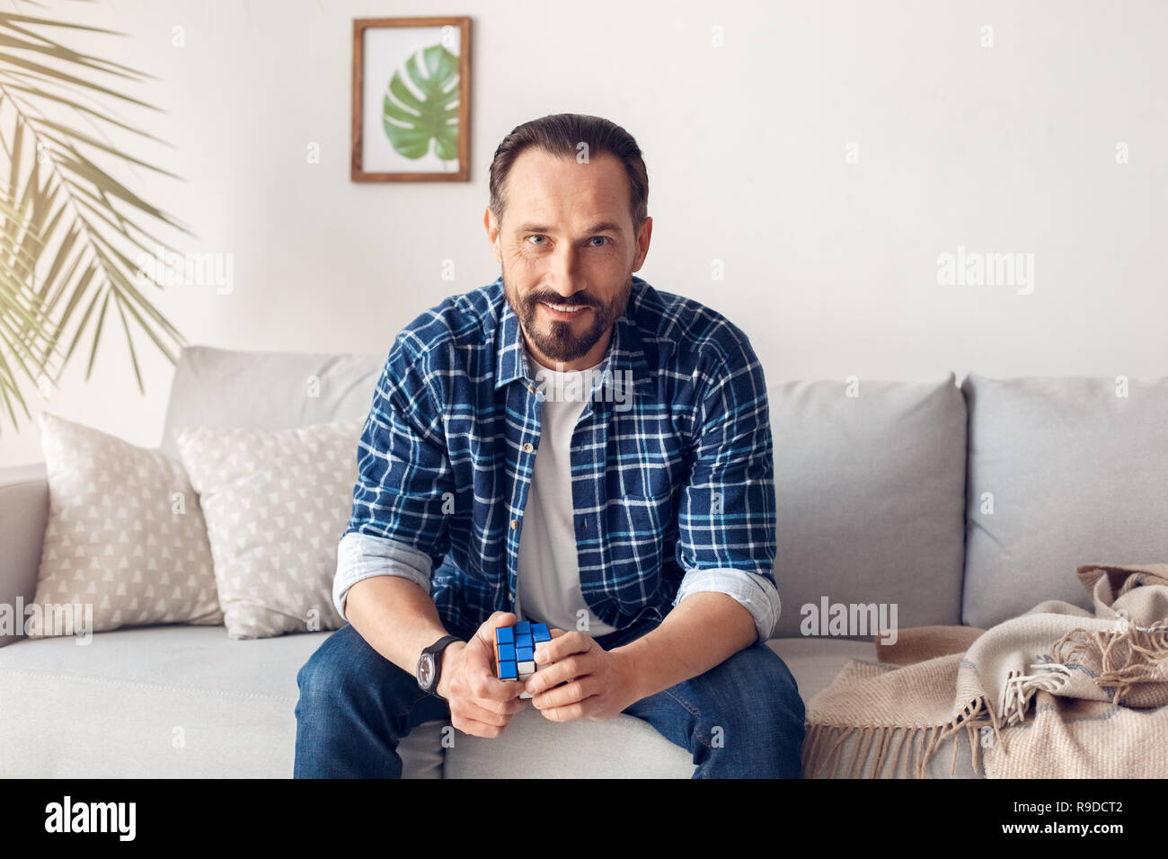 Uomo a casa da solo seduto sul divano tenendo il cubo di Rubik cercando fotocamera a sorridere allegro Foto Stock