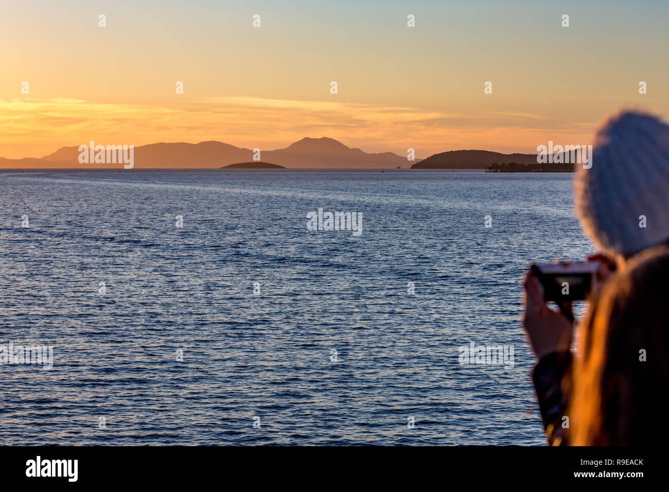 Scenario sunset seascape nel Mar Ionio con la gamma della montagna lontano in background e sfocata irriconoscibile ragazza di fronte con il cappello bianco tenendo il ph Foto Stock