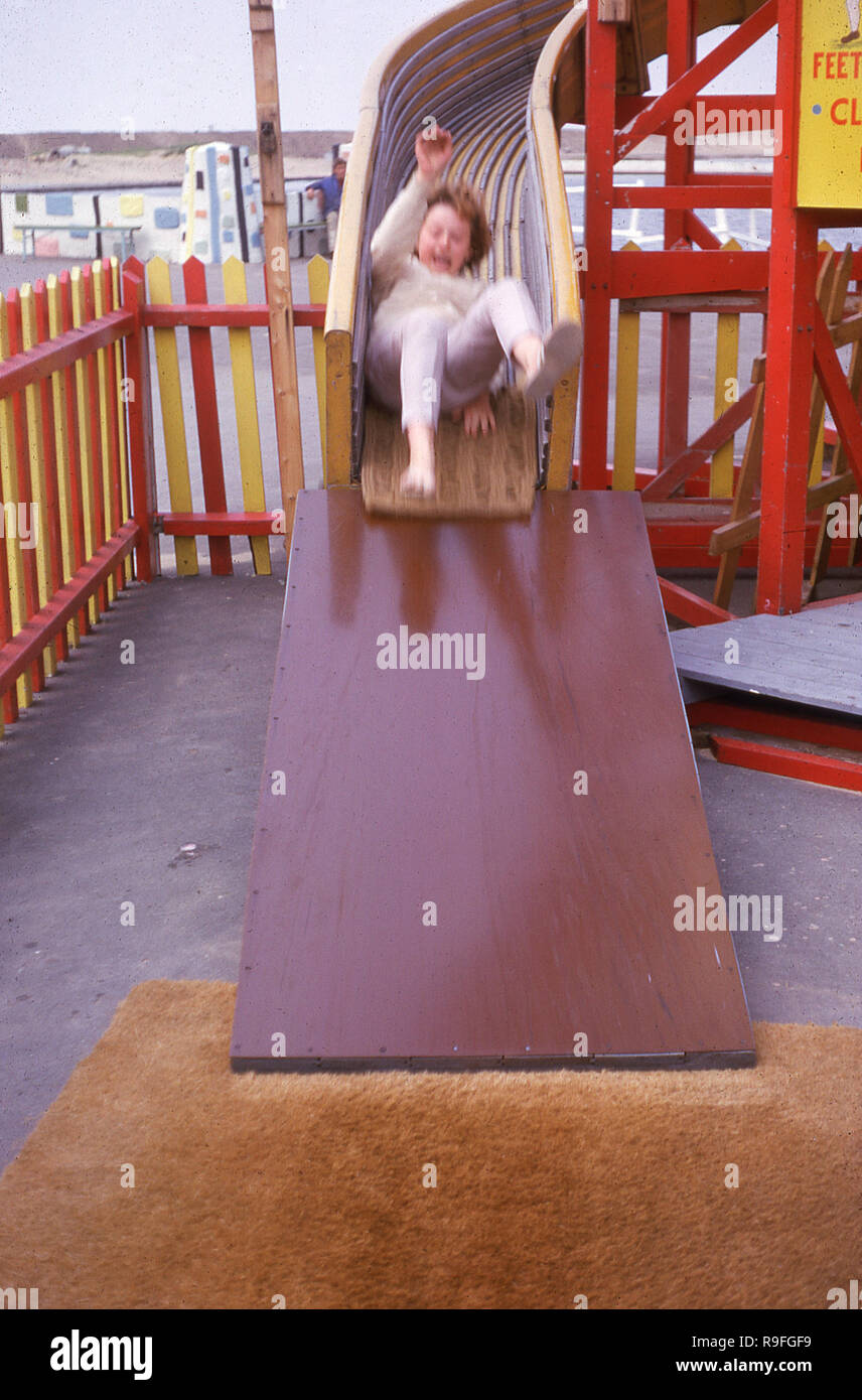 Anni Settanta, una giovane ragazza sulla sua schiena scendendo alla fine di un di legno Helter Skelter scivolo a spirale esterno in un luna park, Inghilterra, Regno Unito. Foto Stock