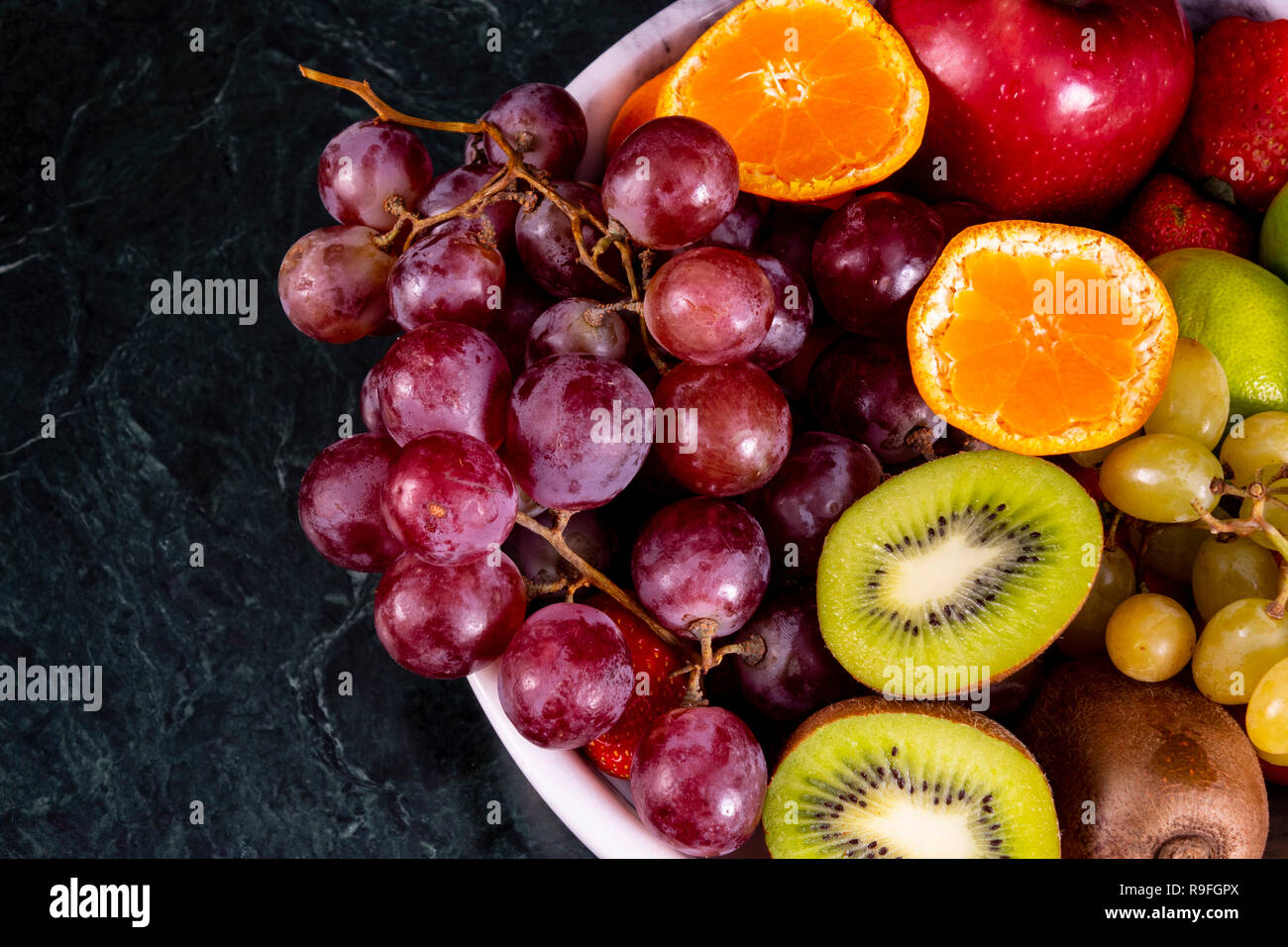 Fresco tropicale fruitson nella ciotola sul marmo nero lo sfondo. Ananas, papaya, mango, kiwi, uva, granato, tangerini, banane, mele, anguria sli Foto Stock