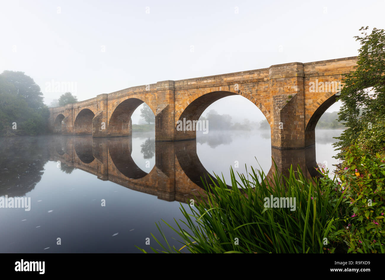 Un ponte di pietra si riflette nell'acqua liscia del fiume che attraversa. Si tratta di sunrise e la nebbia è appeso sopra la superficie del fiume. Foto Stock Un ponte di pietra si riflette nell'acqua liscia del fiume che attraversa. Si tratta di sunrise e la nebbia è appeso sopra la superficie del fiume. Foto Stock