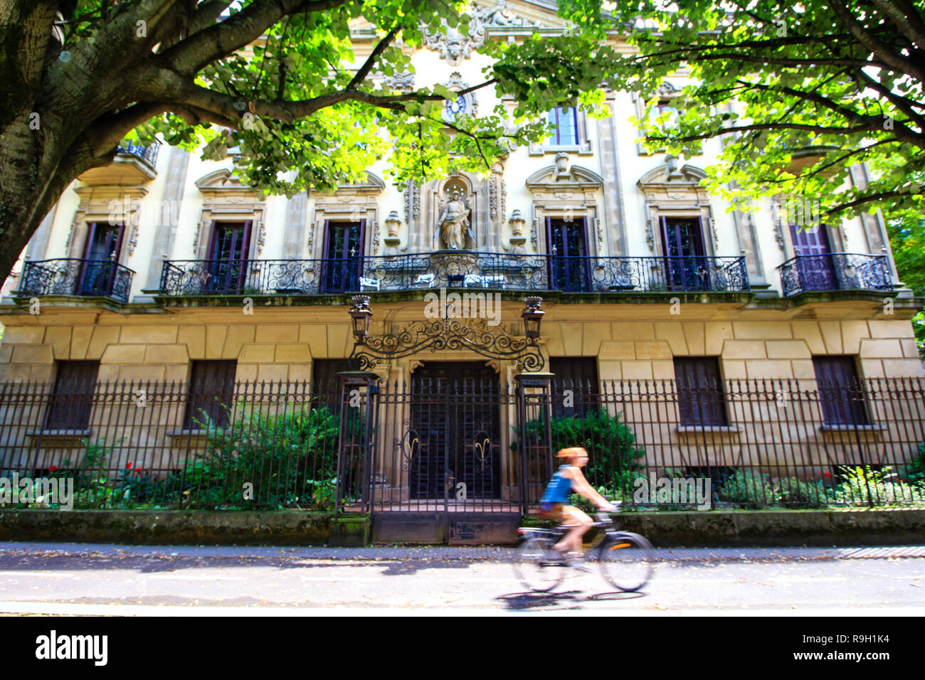 Street scene da San Sebastian in Spagna - ragazza irriconoscibile in bicicletta passa da una bella facciata vecchio edificio sotto alberi verdi. Foto Stock