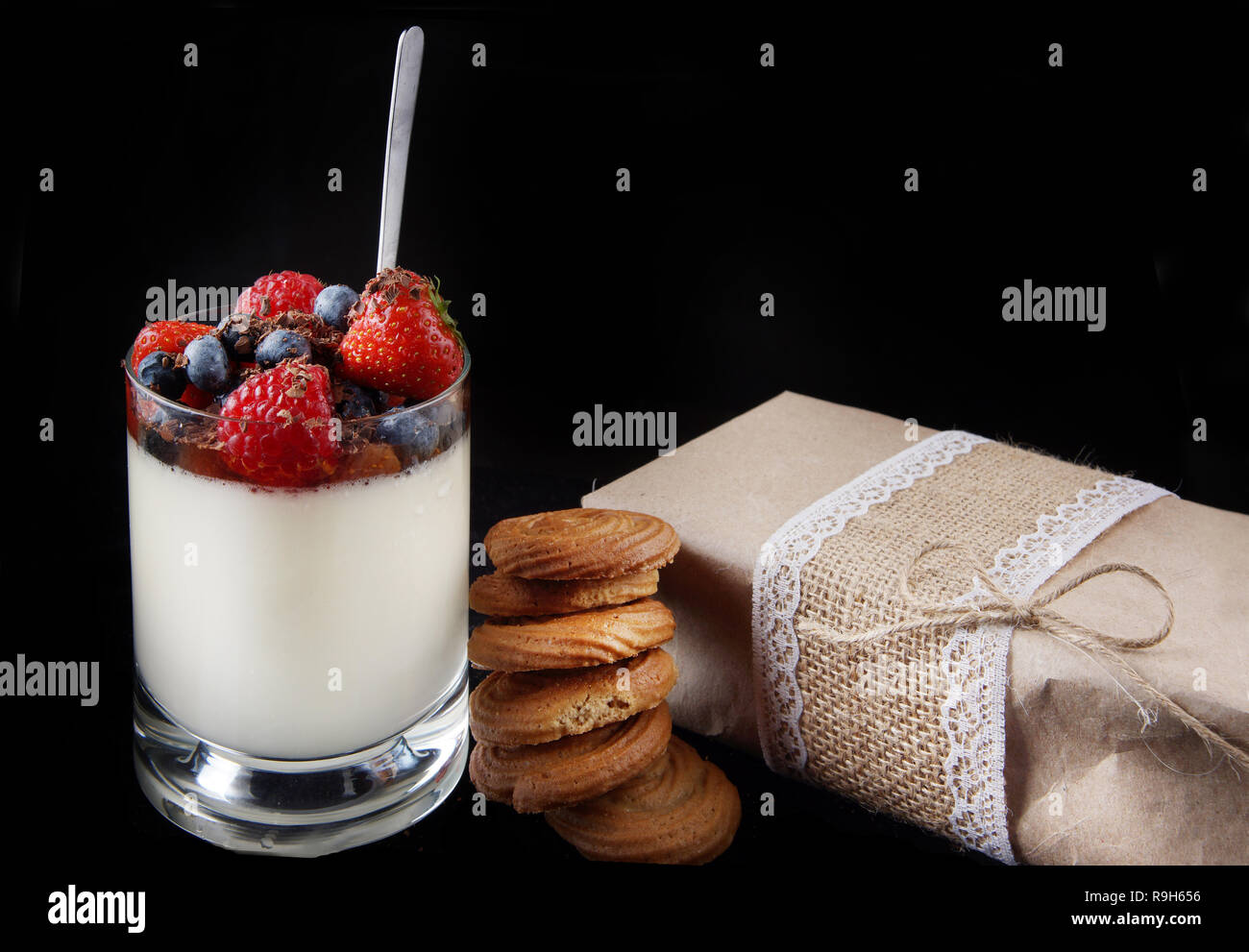 Panakota cremoso dessert italiano in un bicchiere di vetro con frutti di bosco, accanto a una pila di biscotti allo zenzero e un regalo in carta artigianale in stile rustico. Foto Stock