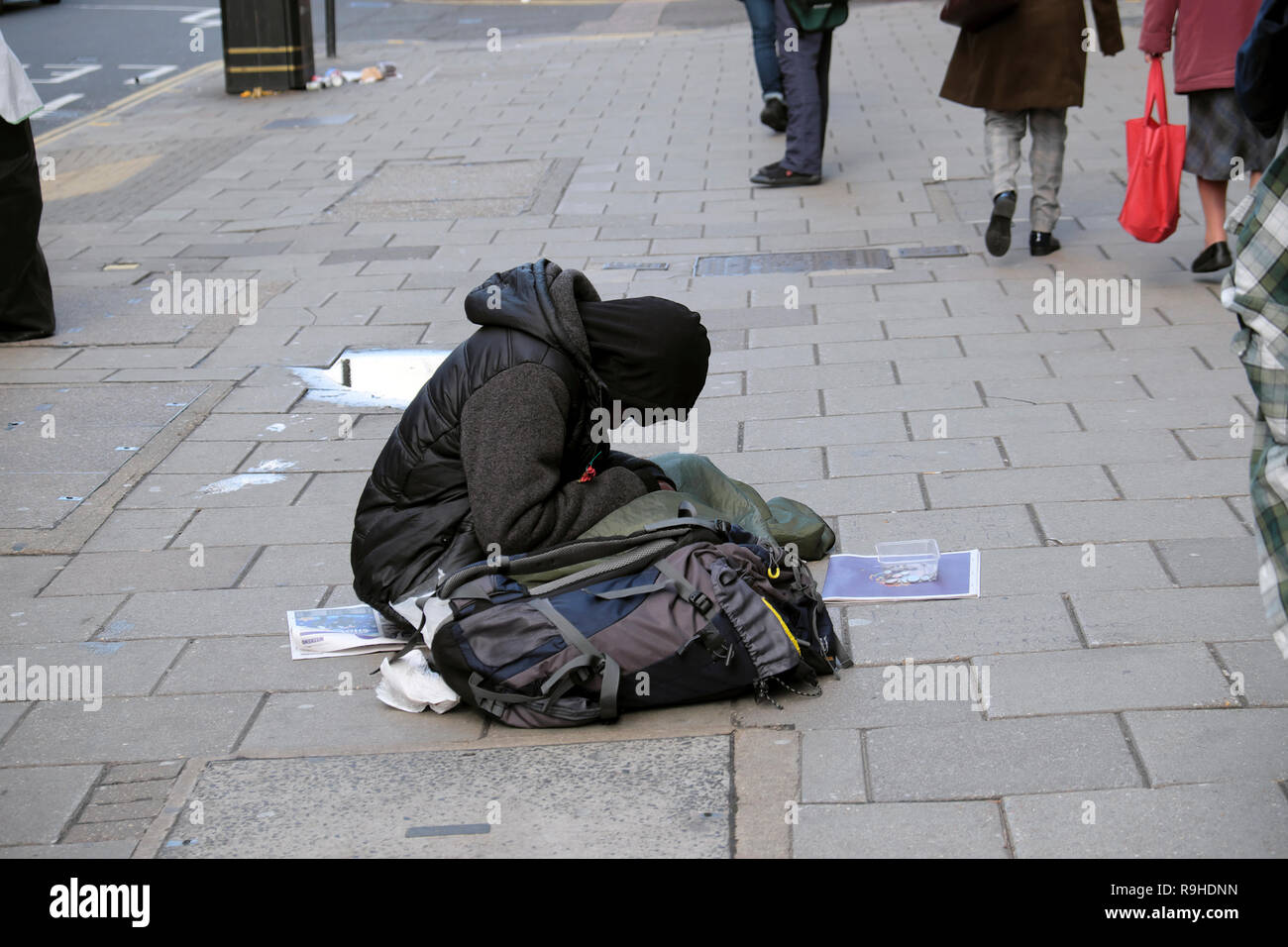 Un senzatetto uomo vestito di nero abbigliamento invernale seduta sul marciapiede marciapiede con testa piegata e la gente a piedi dal London UK Europa KATHY DEWITT Foto Stock