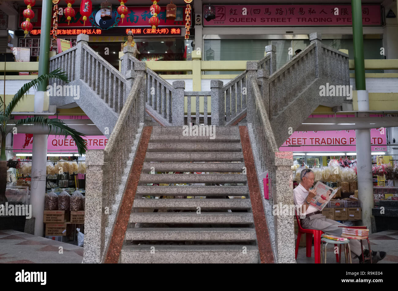 Un lettore di un giornale di Singapore seduto sotto la scala di un piccolo complesso per lo shopping a Waterloo Street, Bugis, Singapore Foto Stock