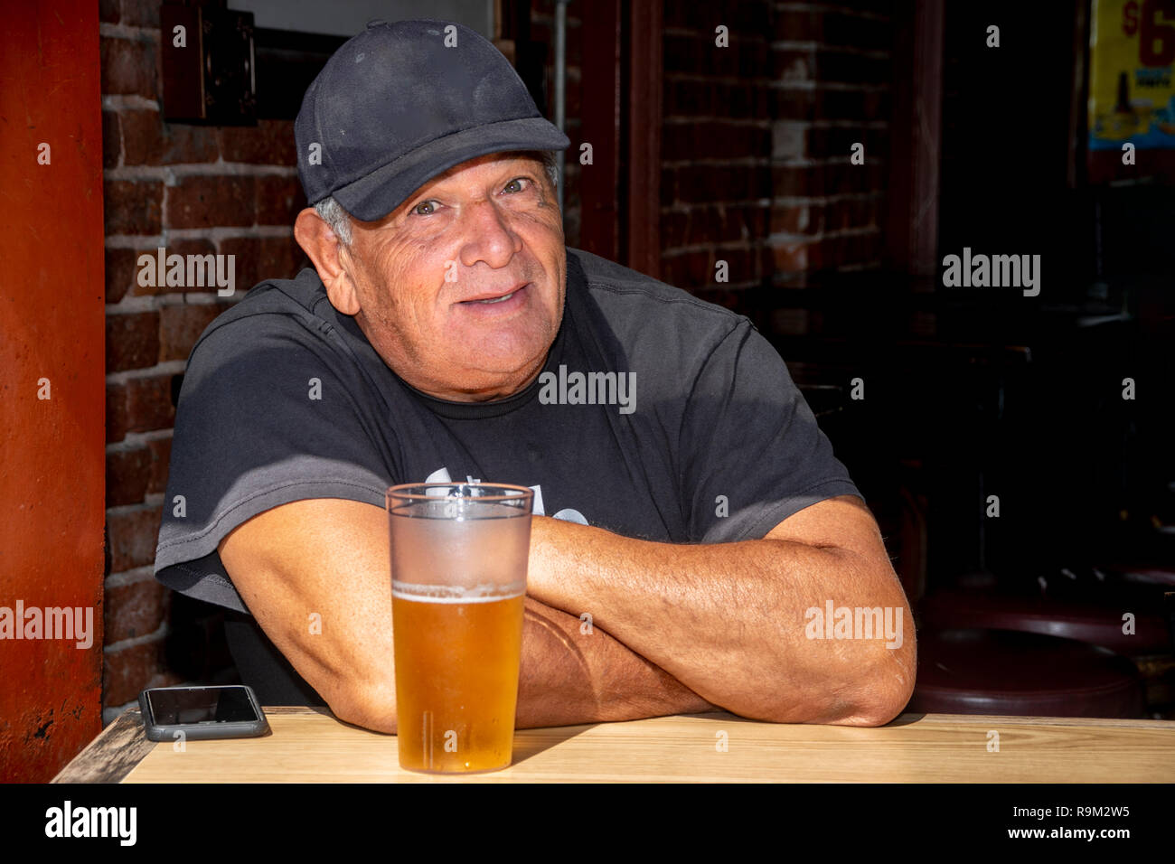 Una sostanziosa senior man in black cap e T shirt gode di una birra alla spina nella finestra di Huntington Beach, CA, taverna su una calda mattina d'estate. Foto Stock