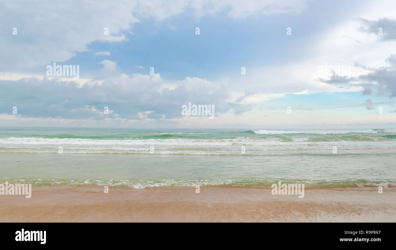 Una vista frontale di una spiaggia di sabbia e un mare verde con piccole onde con un cielo blu con alcune nuvole in background Foto Stock