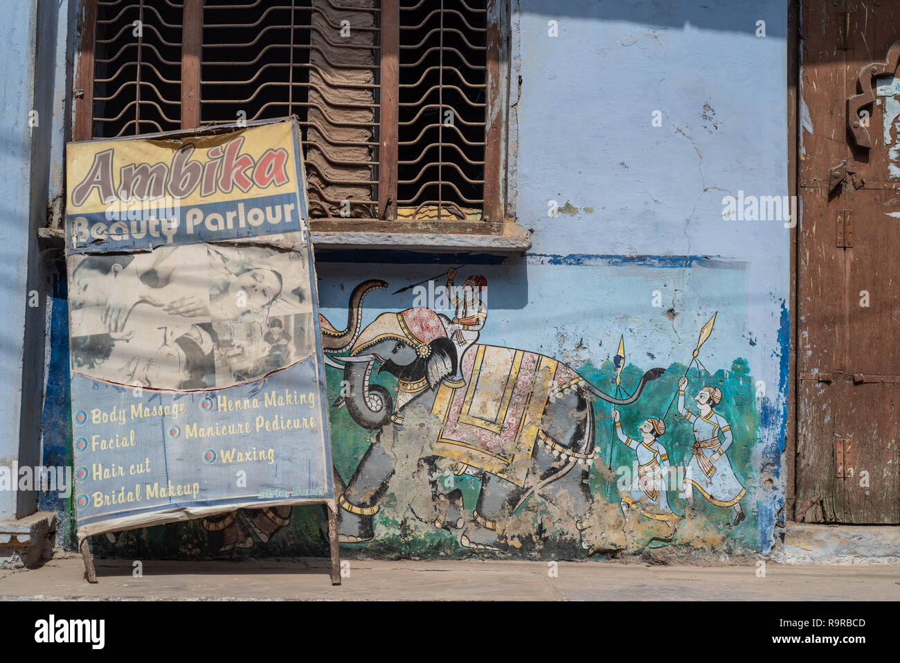 Scruffy, sbiadito e wonky salone di bellezza una scheda sostennero contro un tradizionalmente parete dipinta con una scena di combattimento con gli elefanti. Bundi, Rajasthan Foto Stock