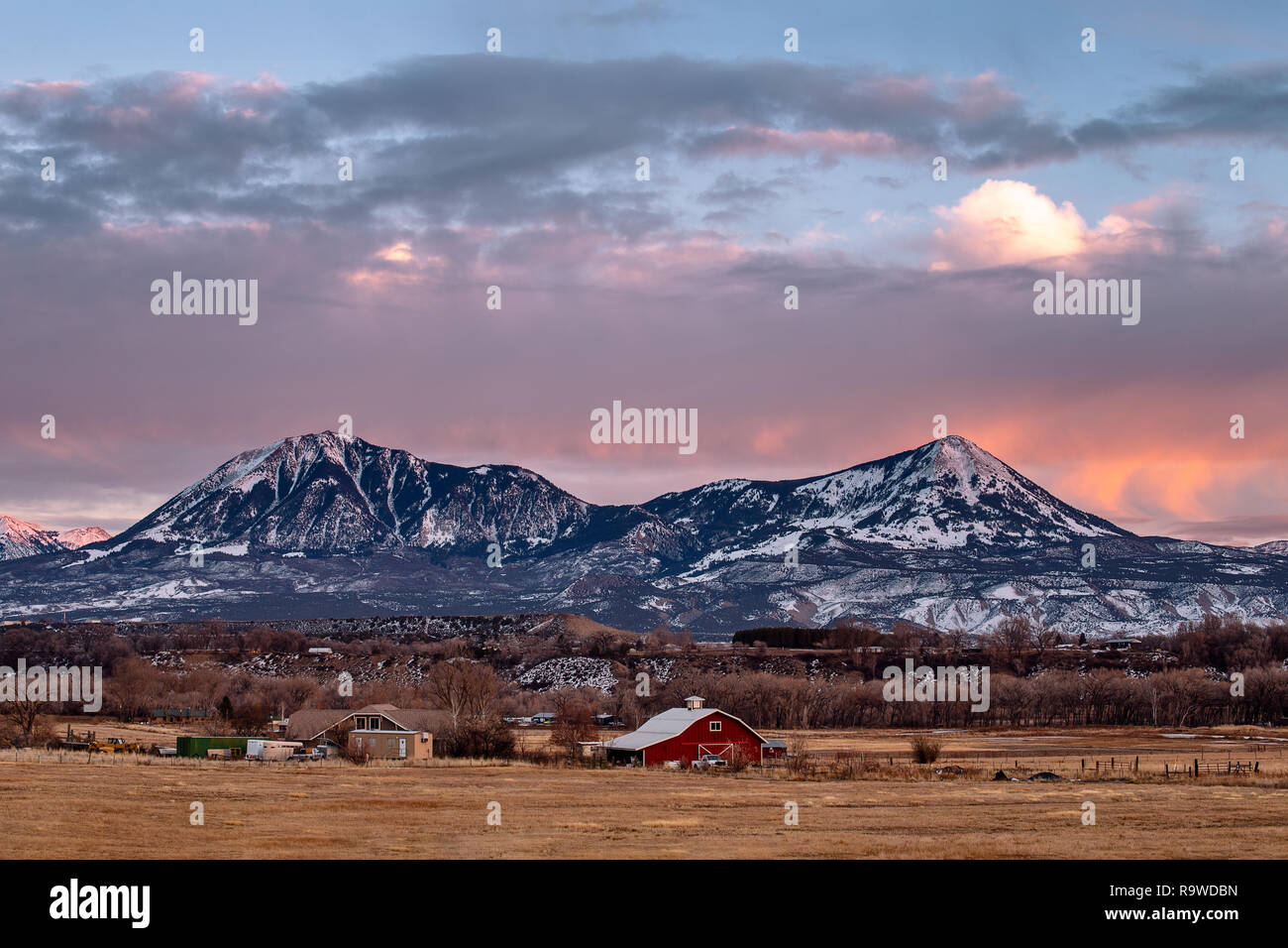 Un tramonto invernale sopra una fattoria lontana e montagne innevate a Delta, Colorado Foto Stock