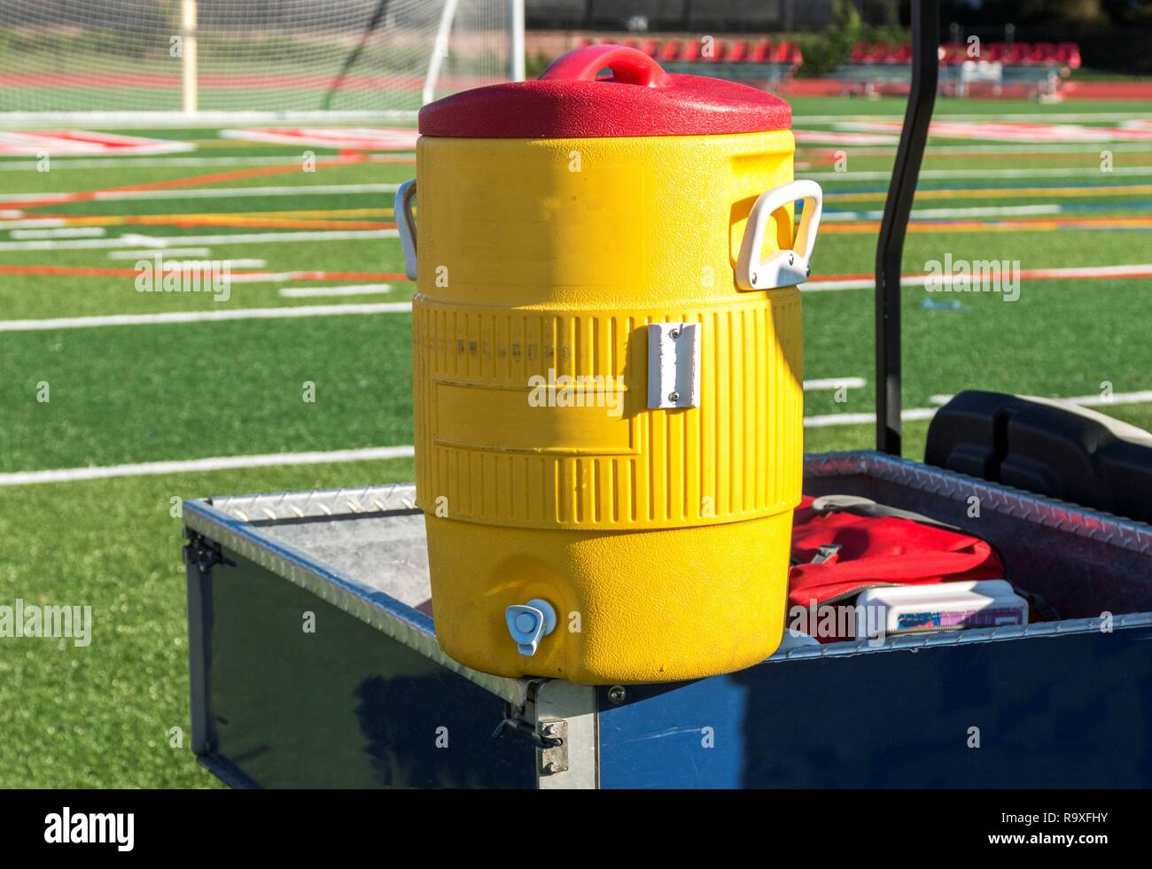 Un giallo dello scambiatore di calore acqua siede su un blu carrello da golf durante una partita di calcio in una scuola locale su un luminoso pomeriggio di sole. Foto Stock