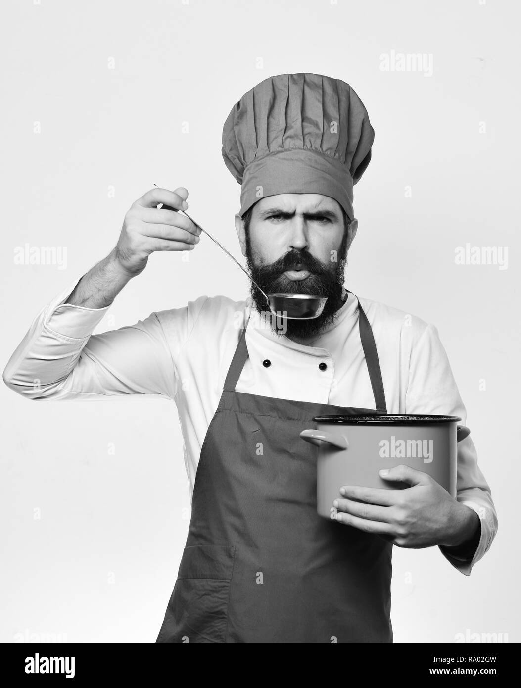 Cucinare con un rigoroso volto in Borgogna è uniforme casseruola con siviera. Cucina professionale concetto. Chef zuppa di gusti o di frutta sciroppata. Uomo con barba detiene pot rosso su sfondo bianco. Foto Stock