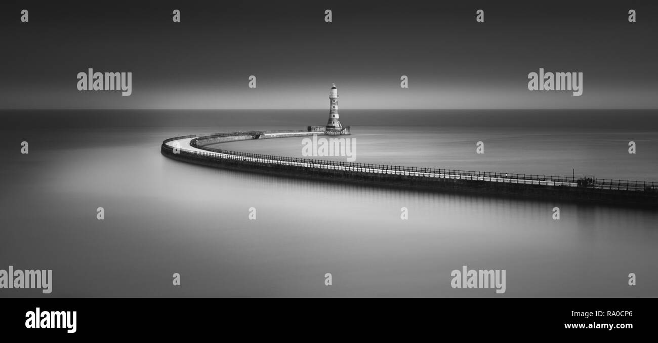 Un moody Arte in bianco e nero lunga esposizione di Roker Pier al tramonto con il lungo percorso di spazzamento dando un soft specchio di riflessione nel Mare del Nord Foto Stock