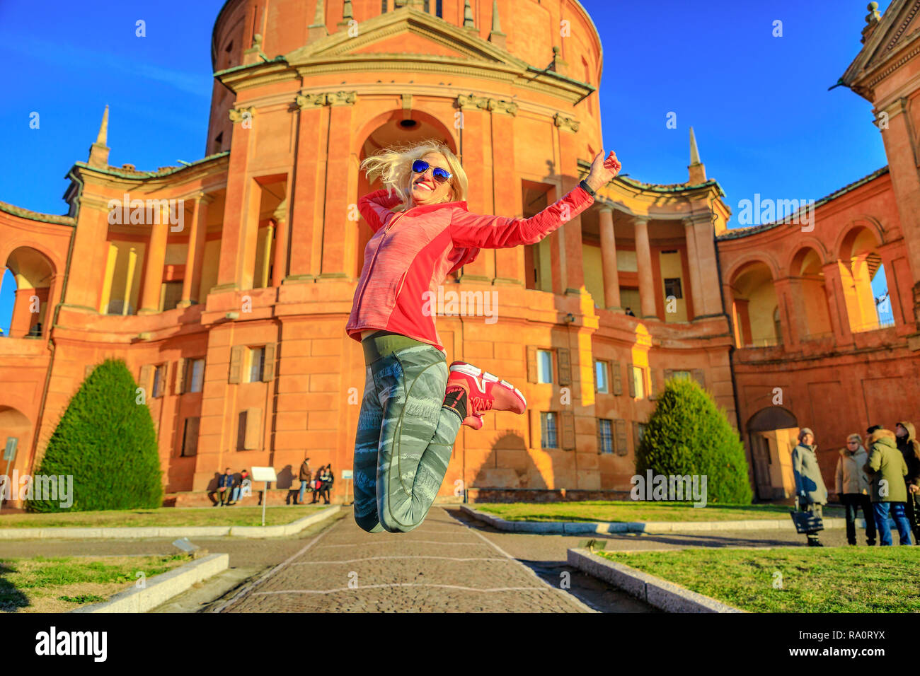Donna felice jumping a Madonna di San Luca Santuario. Attrazioni famose a Bologna, Italia. Femmina bionda godendo della storica Basilica alla luce del tramonto. Soleggiato con cielo blu. Foto Stock