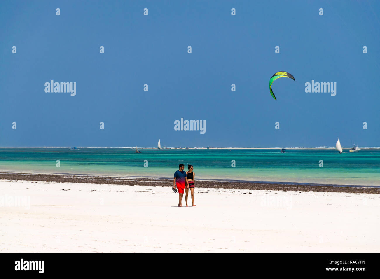 Due turisti a piedi sulla tropicale di sabbia bianca spiaggia di Diani in inizio di mattina di sole con kitesurfisti nell'oceano sullo sfondo, Diani, Kenya Foto Stock