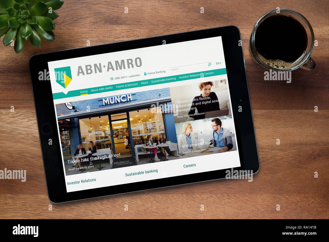 Il sito web di ABN AMRO Bank è visto su un tablet iPad, su di un tavolo di legno lungo con un caffè espresso e una pianta di casa (solo uso editoriale). Foto Stock