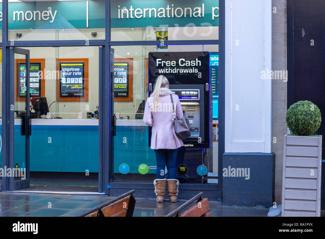 Una persona in un cappotto rosa con lunghi capelli biondi indossa suede pelliccia stivali inverno utilizzando una macchina ATM impostato nella vetrina di un negozio eurochange nella vasca da bagno Foto Stock