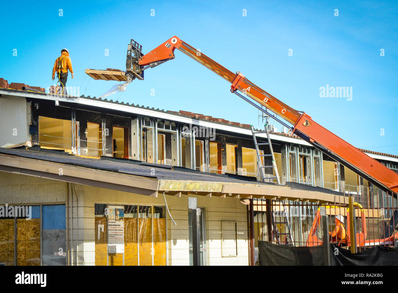 Vista di un lavoratore edile che indossa un elmetto passeggiate sul tetto lungo con gru offrendo un pallet di piastrelle in argilla tetto per rimodellare su un vecchio motel Foto Stock