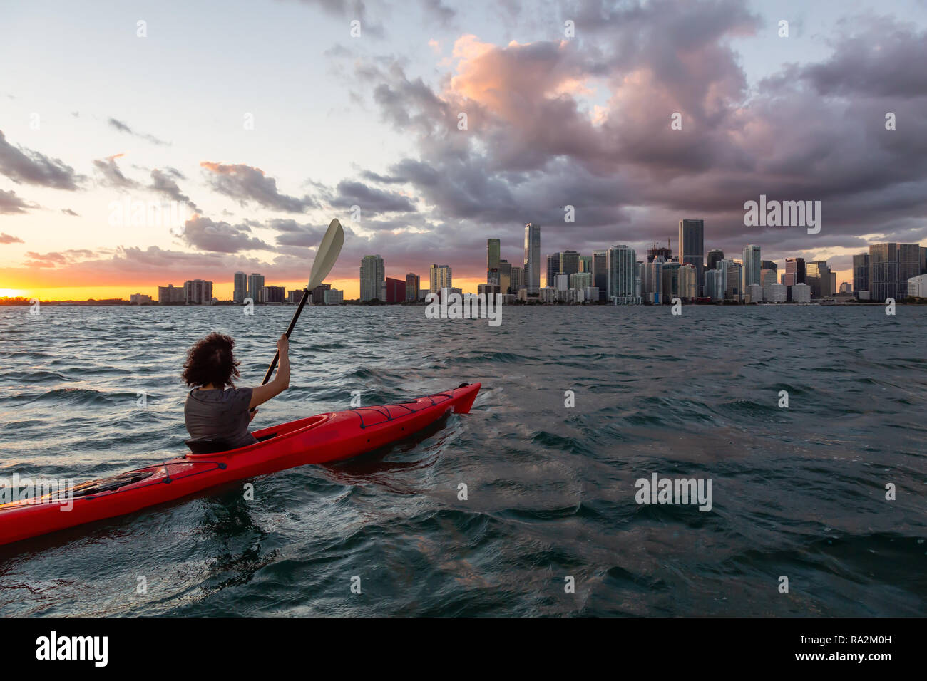 Ragazza avventurosa kayak nella parte anteriore di un moderno centro di Cityscape durante un tramonto spettacolare. Prese a Miami, Florida, Stati Uniti d'America. Foto Stock