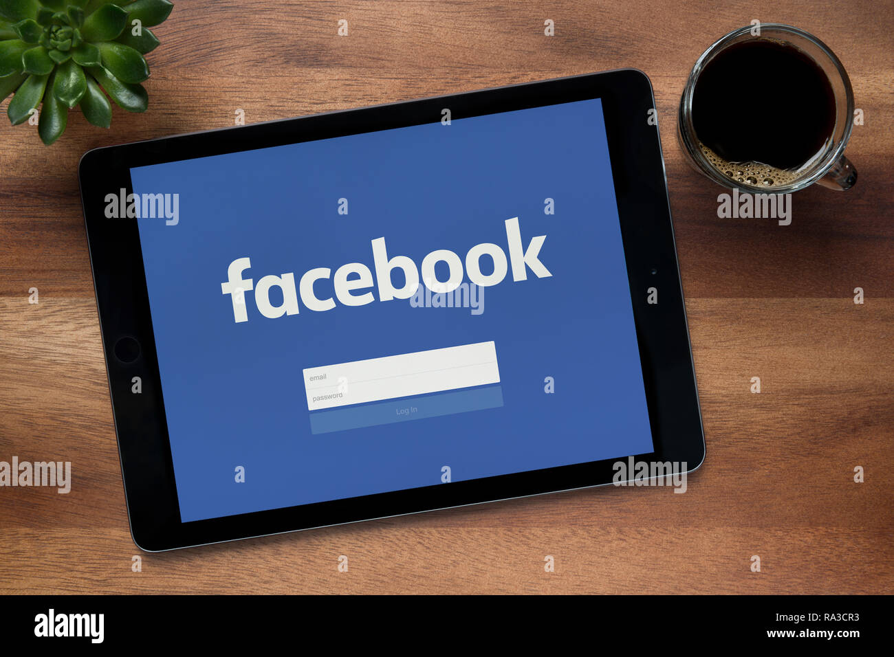 Facebook è visto su un tablet iPad, su di un tavolo di legno lungo con un caffè espresso e una pianta di casa (solo uso editoriale). Foto Stock