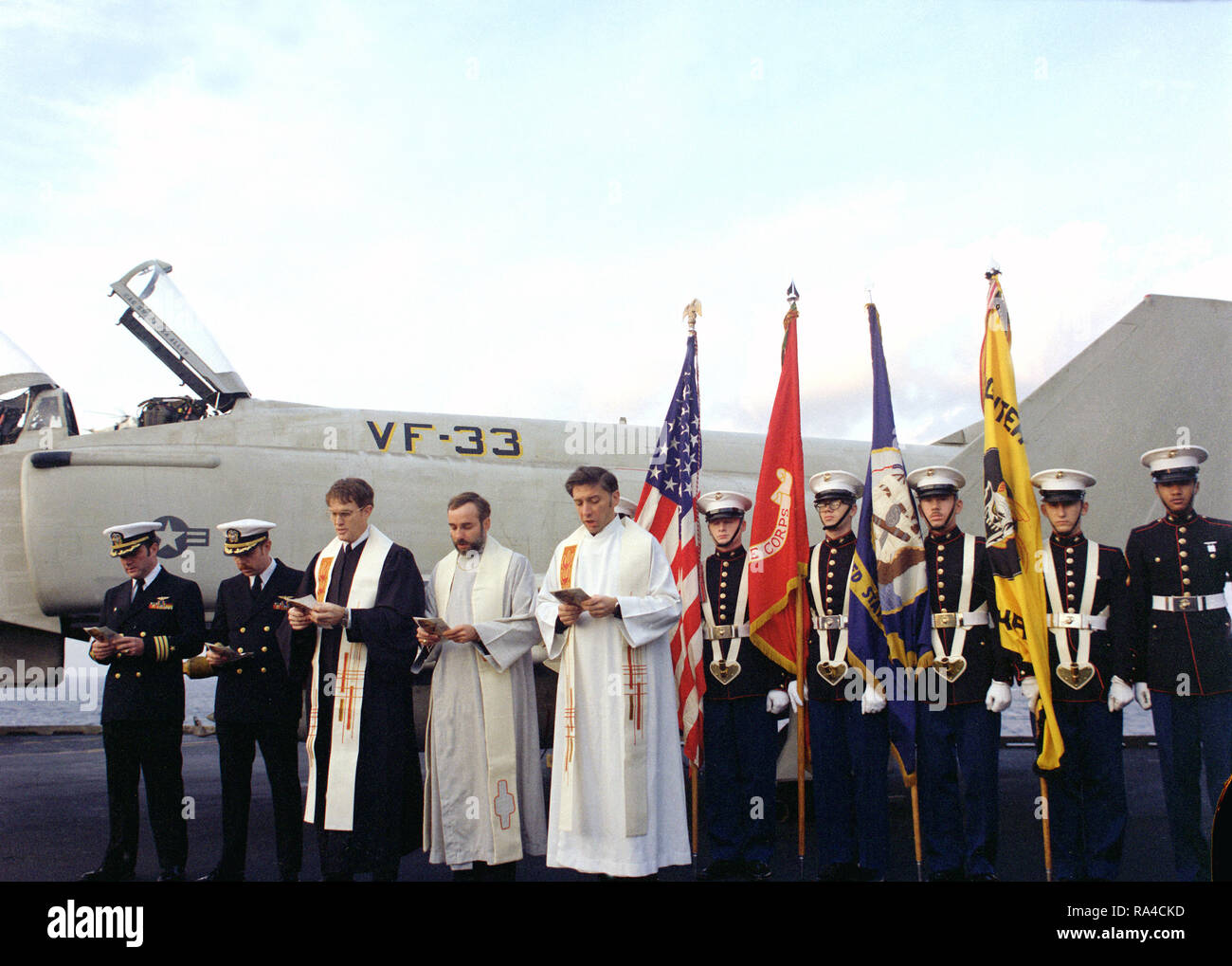 1979 - ufficiali, ecclesiastici e un U.S. Marine Corps color guard stand nei pressi di un aereo di combattimento aereo durante un memoriale di servizio sul ponte di volo della portaerei USS INDIPENDENZA (CV-62). Foto Stock