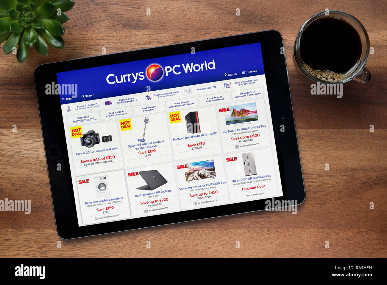 Il sito web di Currys mondo PC è visto su un tablet iPad, su di un tavolo di legno lungo con un caffè espresso e una pianta di casa (solo uso editoriale). Foto Stock