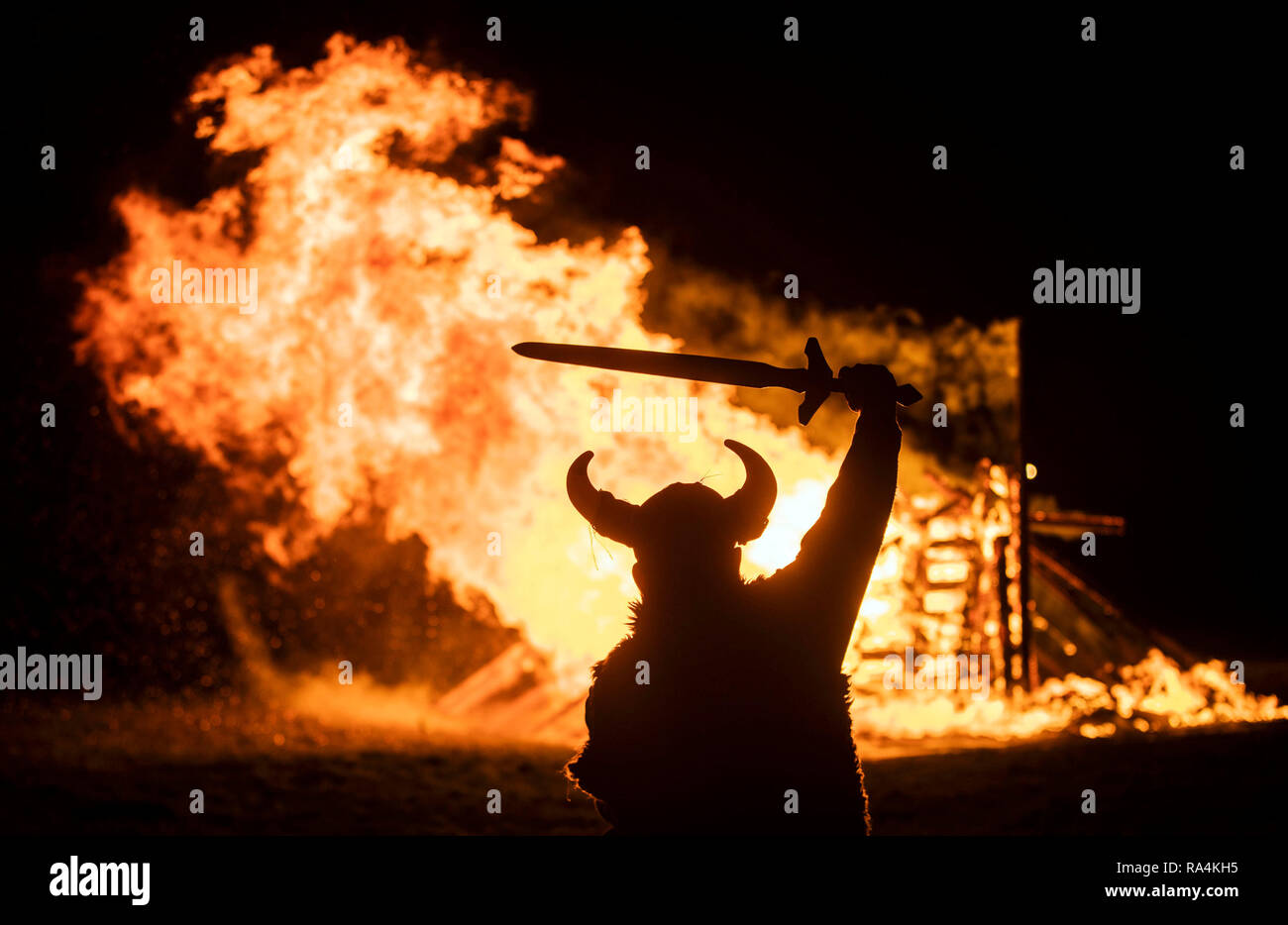 Una persona in costume vichingo di fronte un ardente nave vichinga durante il fuoco Flamborough Festival, Viking parata a tema, tenutasi alla vigilia dell'anno nuovo, a Flamborough vicino a Bridlington, nello Yorkshire, in aiuto degli enti di beneficenza e gruppi delle comunità locali. Foto Stock