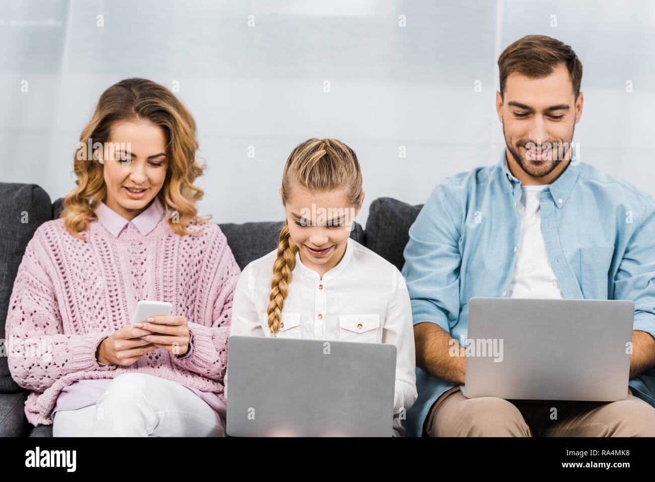 Bella donna seduta sul divano e utilizza lo smartphone durante la figlia e suo marito la digitazione su tastiere di computer portatile in salotto Foto Stock