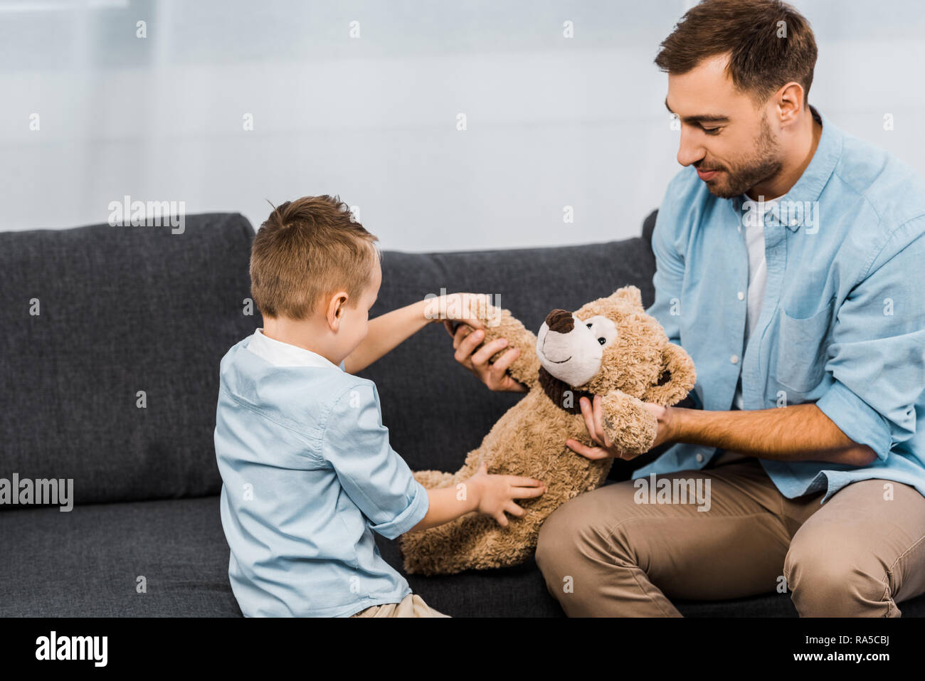 Sorridente padre e figlio seduti sul divano e tenendo orsacchiotto in salotto Foto Stock