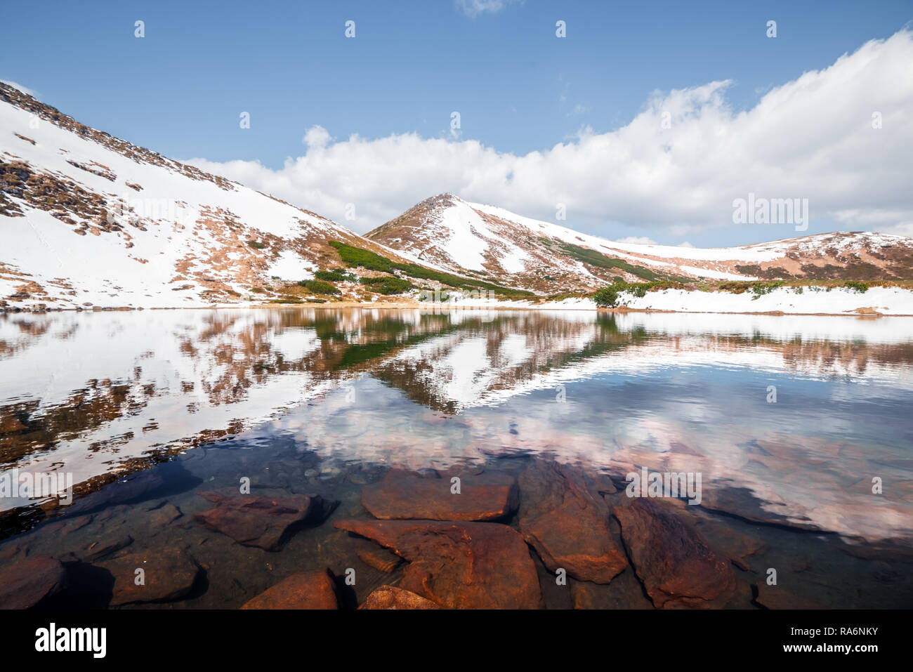Molla di lago di montagna con acqua limpida e pietre rosse. Il pittoresco paesaggio invernale con colline innevate sotto un cielo blu Foto Stock