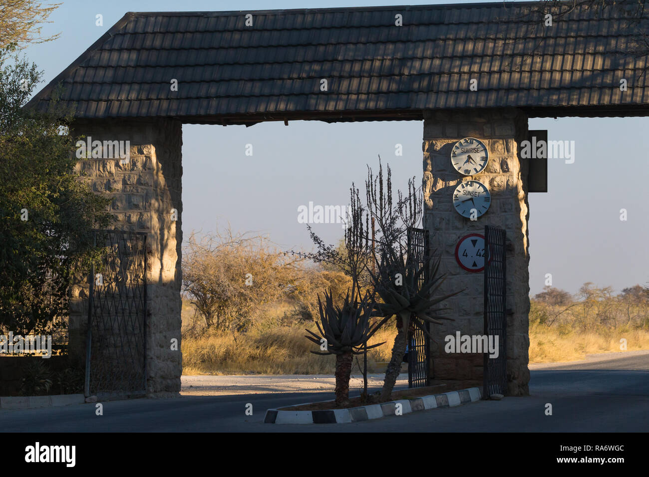 Okaukuejo rest camp cancello aperto e dando tempi di chiusura e di apertura su un orologio sul muro di pietra nel Parco Nazionale Etosha, Namibia Foto Stock