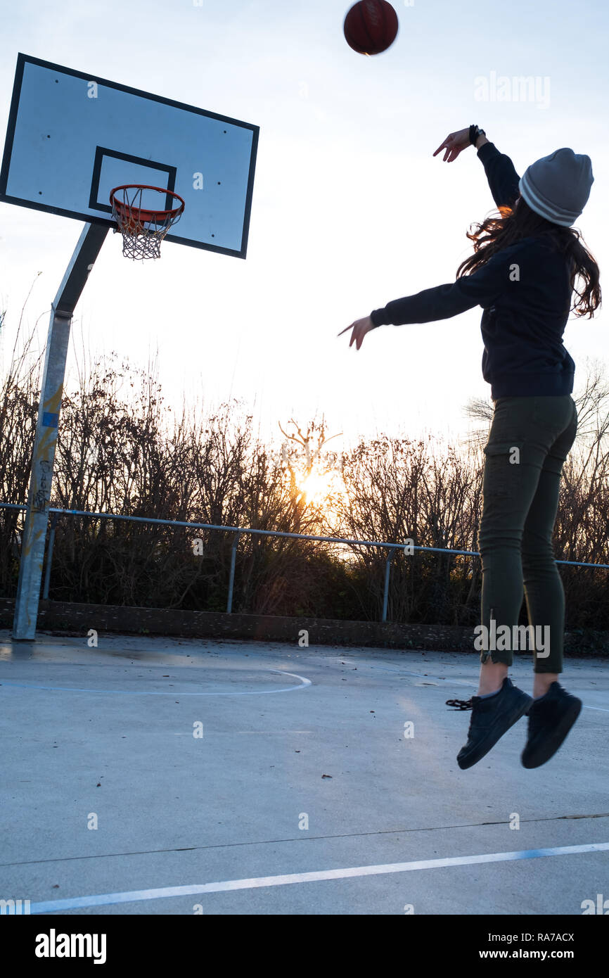 Young Teen girl, metà aria, tiro jump shot sul campo di pallacanestro, retroilluminato al tramonto, composizione verticale, spazio di copia Foto Stock
