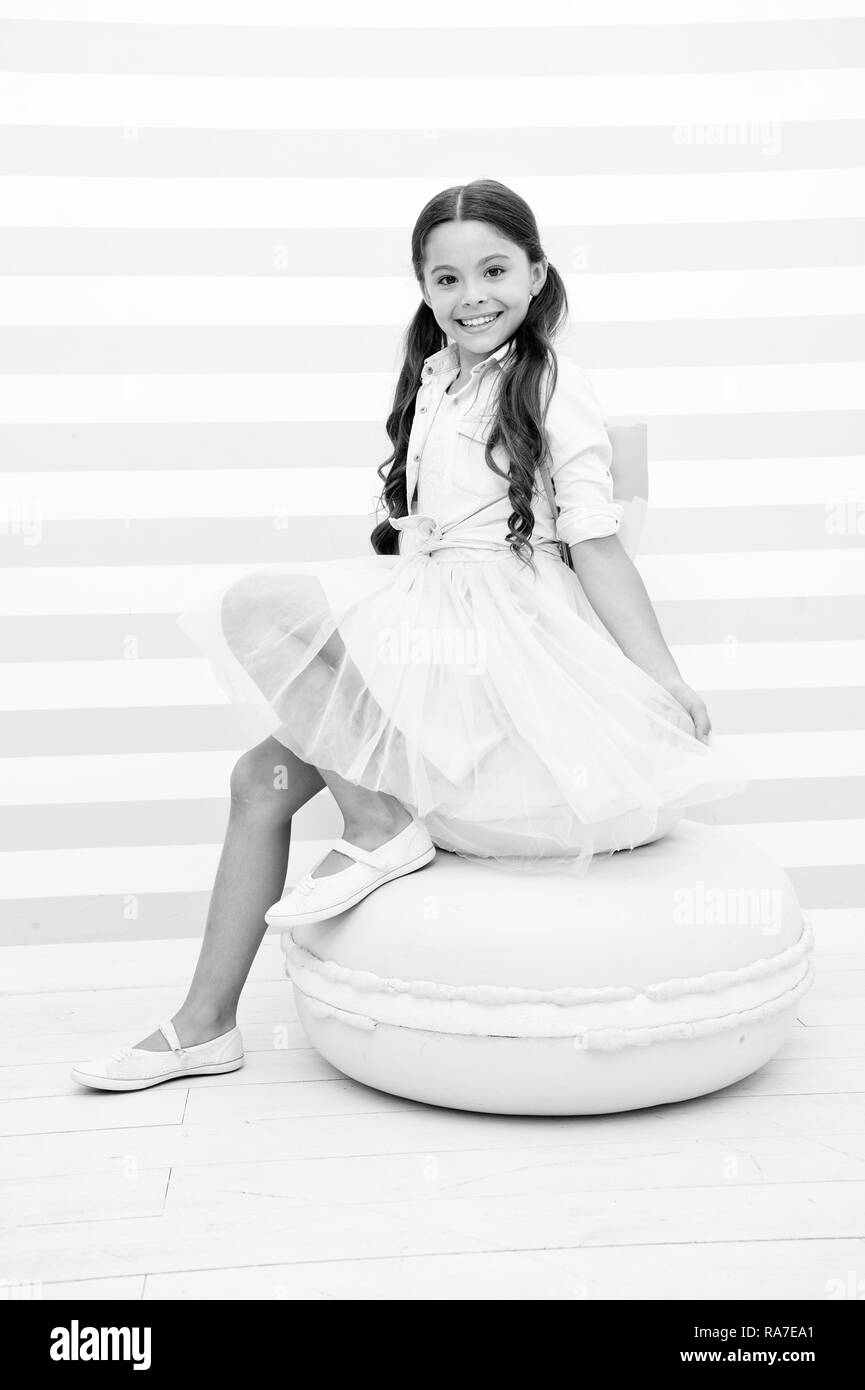 Dolce principessa. Bambino adorabile ragazza sorridente in posa sulla cute amaretto dolce pouf. Kid lunghi capelli ricci indossare abiti alla moda. Ragazza con grazioso sacchetto ponendo sullo sfondo a strisce. Le piace essere un modello. Foto Stock