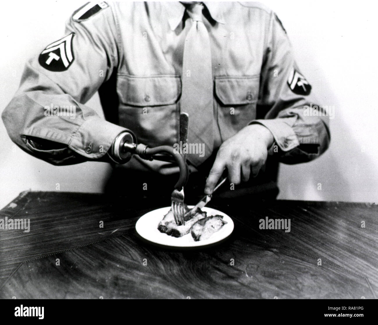Vista di un uomo in uniforme militare rappresentata dal collo in giù seduti ad un tavolo. L'uomo detiene una forcella nella sua protesico gancio destro e con la sua mano sinistra taglia un pezzo di carne sul piatto. Foto Stock