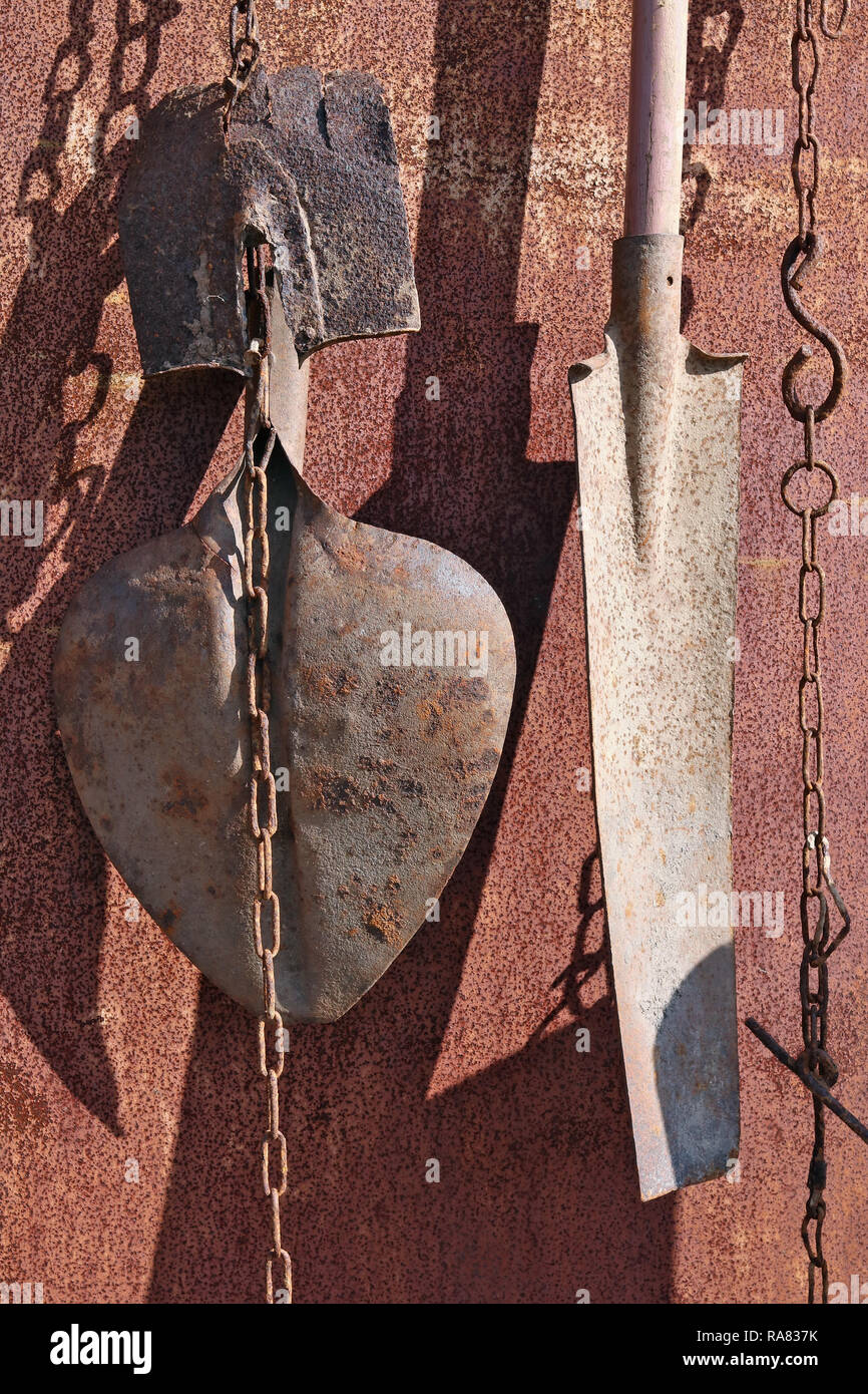 Sul metallo arrugginito parete del capannone di villaggio appendere su catene utilizzate vecchie vintage pale agricoli. Soleggiata giornata di primavera shot Foto Stock