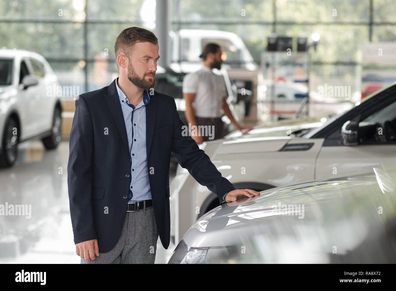 Gli uomini alla ricerca di veicoli in car showroom. Uomo Barbuto che  indossa in dark blue Jacket guardando auto e tenendo una mano sul cofano  per auto. Persone osservando le automobili per