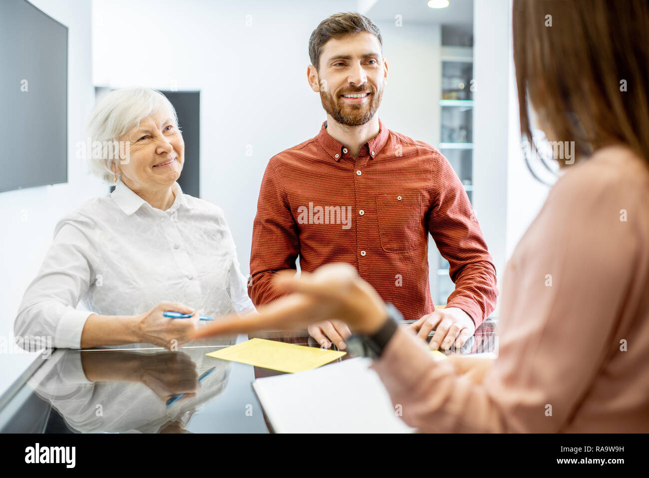 Uomo sorridente con il suo senior madre parlando con la receptionist in ospedale Foto Stock