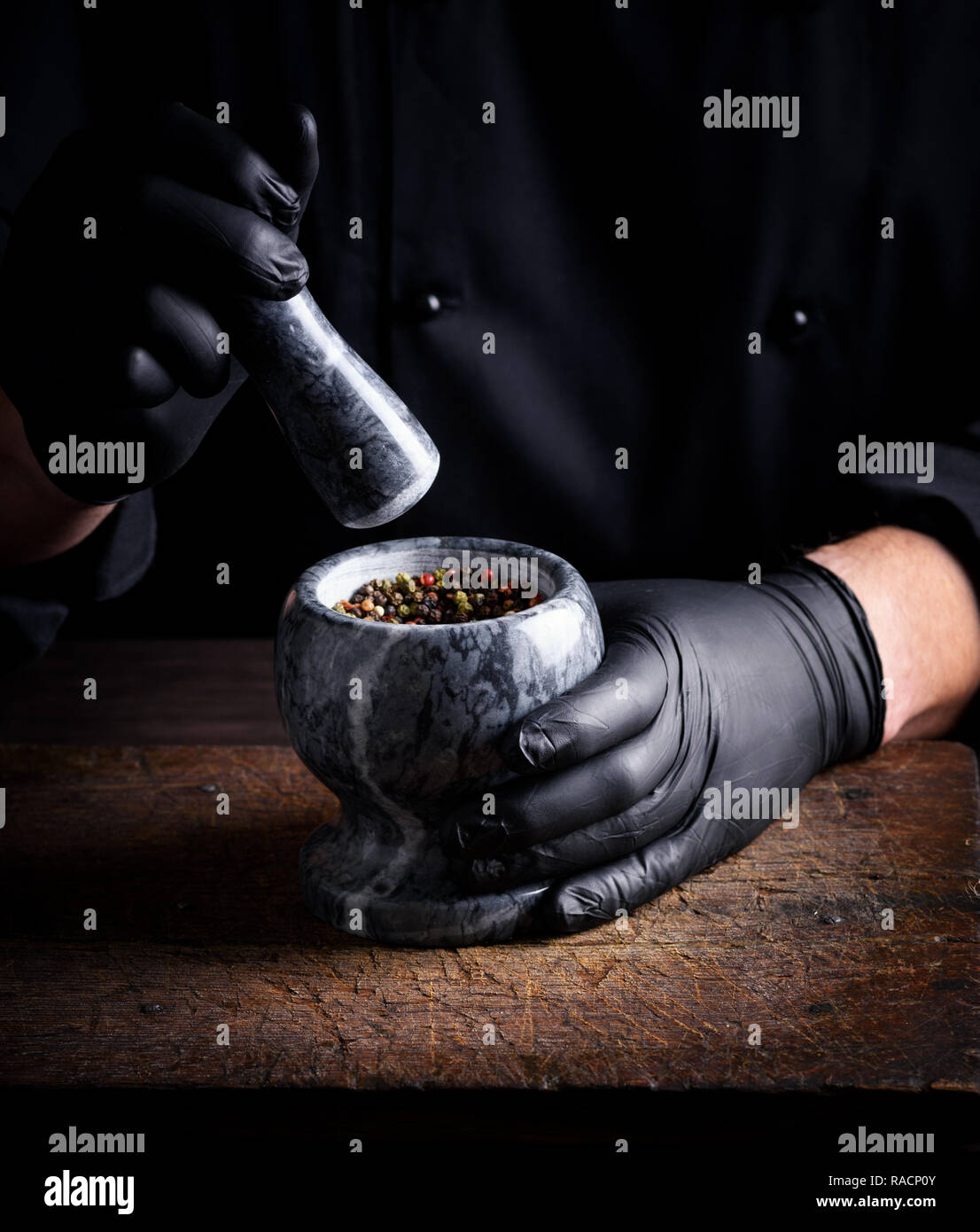 Chef in nero Guanti in lattice detiene un mortaio di pietra con una miscela di peperoni, cottura e macinazione di spezie Foto Stock