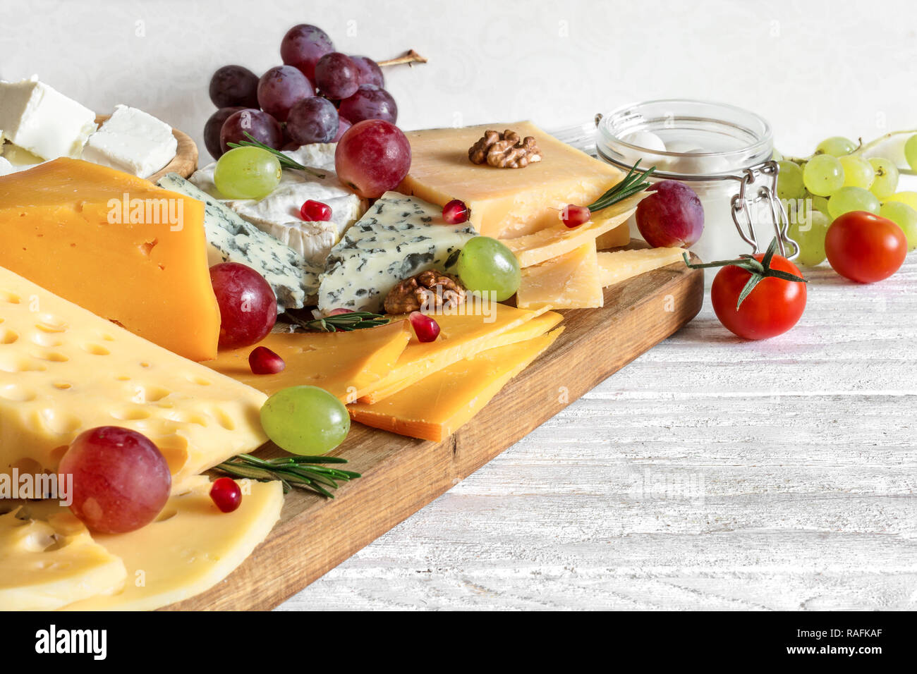 Vari tipi di formaggio servita sul tagliere di legno con uve, melograno, rosmarino e i pomodori ciliegia. close up Foto Stock