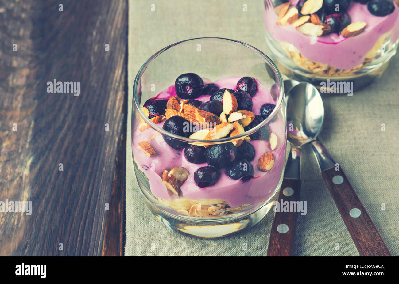 Dolce di farina di avena, banana, yogurt, mirtilli e mandorla in ciotole in vetro su legno rustico sfondo. Vintage nei toni dell'immagine. Foto Stock