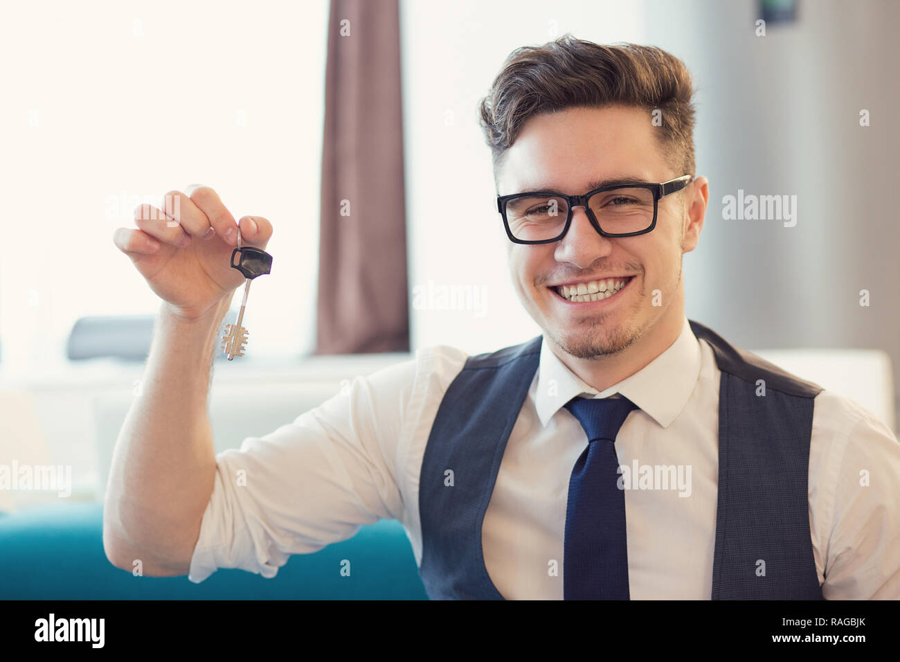 Giovane uomo sorridente in occhiali in possesso della chiave da casa nuova seduta sul lettino Foto Stock