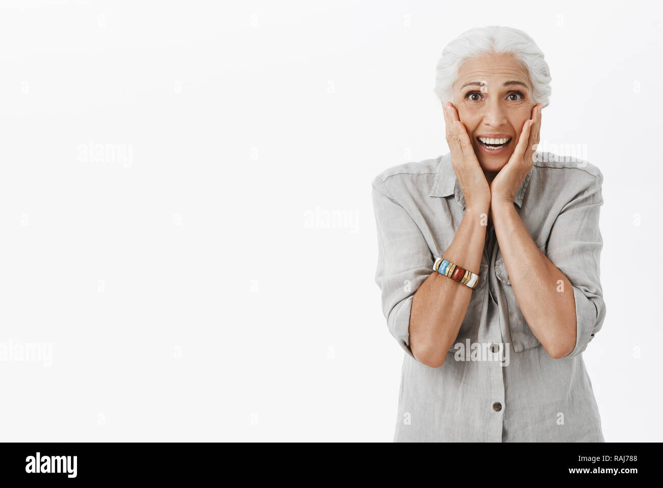 Studio shot di senior donna reagire a sorpresa. Ritratto di toccato e felici carino vecchia signora con i capelli bianchi toccando le guance divertito e sorridente broadlt ricevere regalo bellissimo oltre il muro grigio Foto Stock